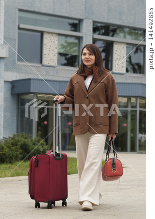 A woman in neutral tones with a brown coat and red suitcase stands outside. The frame conveys elegance, simplicity, and the comfort of travel in style. 141492885
