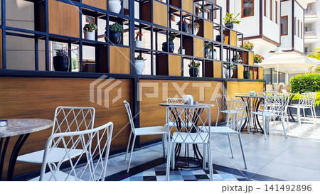 Minimalist caf terrace with empty tables and chairs in a bright hotel courtyard surrounded by plants. The peaceful travel scene reflects vacation relaxation, outdoor dining and sunny resort mornings. 141492896