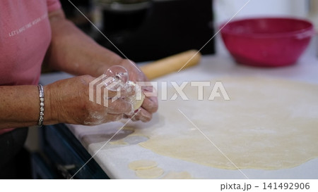 Glass rim cuts multiple round shapes from rolled dough as hands prepare pastry on a kitchen counter. The scene illustrates homemade cooking activity, useful for baking tutorials, food lifestyle Glass rim cuts multiple round shapes from rolled dough as hands prepare pastry on a kitchen counter. The scene illustrates homemade cooking activity, useful for baking tutorials, food lifestyle 141492906