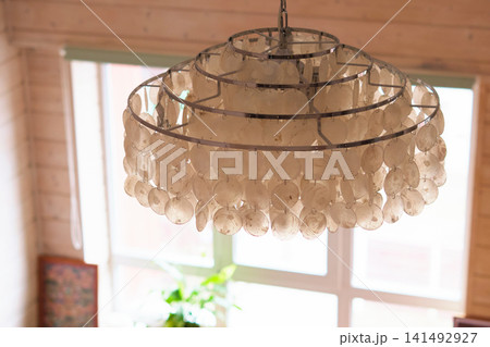Close up of a shell chandelier hanging in front of a bright window inside a warm wooden room. Natural textures and soft daylight bring boho style, home comfort, and relaxed interior design. Close up of a shell chandelier hanging in front of a bright window inside a warm wooden room. Natural textures and soft daylight bring boho style, home comfort, and relaxed interior design. 141492927