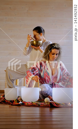 Eye level view of a focused woman playing crystal singing bowls as another creates sound with a Tibetan bowl behind. Shared meditation helps ease tension, improve focus, and restore calm. Eye level view of a focused woman playing crystal singing bowls as another creates sound with a Tibetan bowl behind. Shared meditation helps ease tension, improve focus, and restore calm. 141492932