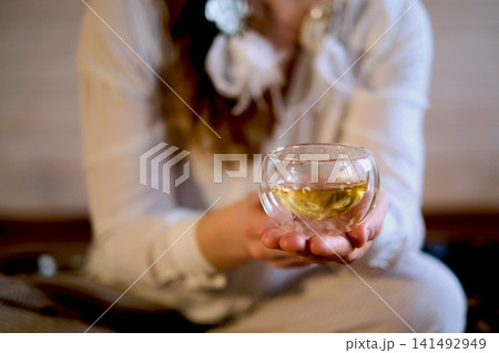 Selective focus shows a glass teacup in the open palm of a woman, with her seated figure softly blurred behind. Tea breaks and self care routines encourage relaxation, balance, and wellbeing. 141492949