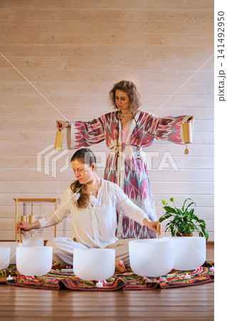 Eye level view of women using crystal bowls and wooden chimes during a candlelit sound therapy ritual on a bright rug. Mindful vibrations bring comfort, grounding, stress relief, and calm. Eye level view of women using crystal bowls and wooden chimes during a candlelit sound therapy ritual on a bright rug. Mindful vibrations bring comfort, grounding, stress relief, and calm. 141492950