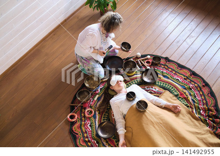 High angle view of a therapist playing a Tibetan singing bowl near a woman resting with an eye pillow on a colorful rug. Private sound bath rituals support deep relaxation, stress relief, and calm. High angle view of a therapist playing a Tibetan singing bowl near a woman resting with an eye pillow on a colorful rug. Private sound bath rituals support deep relaxation, stress relief, and calm. 141492955