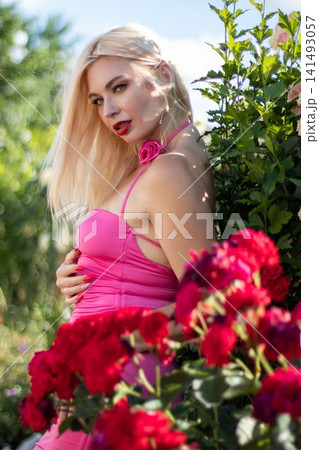 Adult blonde woman poses in profile with one hand on her waist beside vivid red roses. Elegant outdoor portrait for fashion, beauty, and floral advertising. Adult blonde woman poses in profile with one hand on her waist beside vivid red roses. Elegant outdoor portrait for fashion, beauty, and floral advertising. 141493057