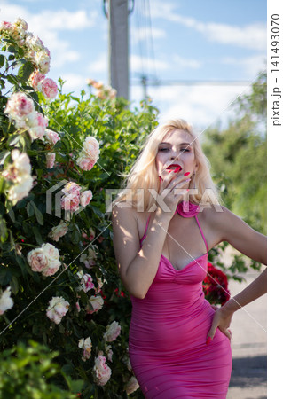 Adult blonde woman touches her lips while posing with one hand on her hip beside blooming roses. Suitable for beauty, makeup, fashion, and glamour content. Adult blonde woman touches her lips while posing with one hand on her hip beside blooming roses. Suitable for beauty, makeup, fashion, and glamour content. 141493070
