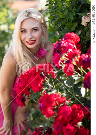 Smiling adult blonde woman crouches beside vivid red roses and looks at the camera in a sunny garden. Suitable for joyful fashion and floral lifestyle themes. Smiling adult blonde woman crouches beside vivid red roses and looks at the camera in a sunny garden. Suitable for joyful fashion and floral lifestyle themes. 141493080