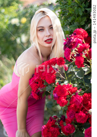Adult blonde woman leans toward a cluster of vivid red roses and looks at the camera. Bright floral portrait for fashion, beauty, and summer lifestyle use. 141493082