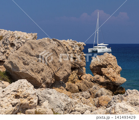 Rocky Coastline with Sailing Catamaran in the Mediterranean Sea near Rhodes, Greece Rocky Coastline with Sailing Catamaran in the Mediterranean Sea near Rhodes, Greece 141493429