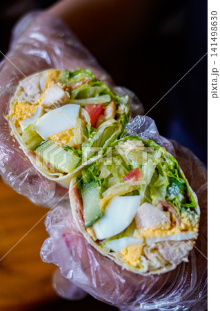 Close-up of tortilla wrap filled with chicken, egg and vegetables held in hand. Healthy food and takeaway concept. Close-up of tortilla wrap filled with chicken, egg and vegetables held in hand. Healthy food and takeaway concept. 141498630