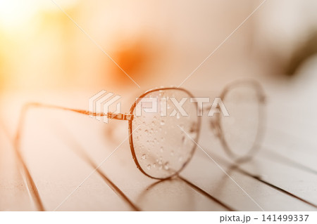 Eyeglasses droplets sunset round gold spectacles resting on wooden table with water drops during warm golden hour light 141499337