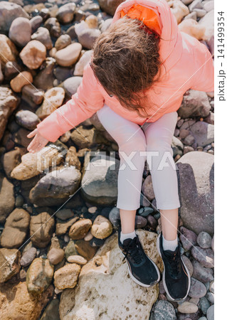 Beach pebbles relaxation girl in bright peach jacket sits on large rocks by the sea coast during sunny vacation day Beach pebbles relaxation girl in bright peach jacket sits on large rocks by the sea coast during sunny vacation day 141499354