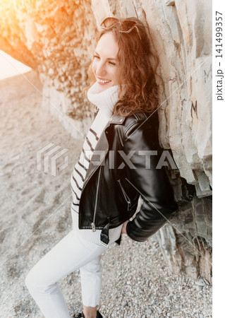 Woman leather jacket coastline smiling while leaning against a rock wall during a golden hour sunset travel escape Woman leather jacket coastline smiling while leaning against a rock wall during a golden hour sunset travel escape 141499357