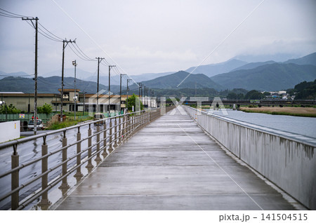 雨が降る曇り空の下で真新しい防潮堤の上の小道と電柱や山並みが奥へ続く風景 雨が降る曇り空の下で真新しい防潮堤の上の小道と電柱や山並みが奥へ続く風景 141504515