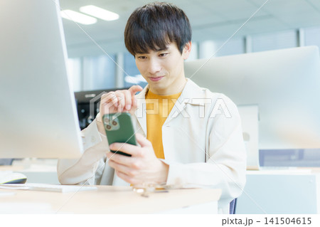 デスクの前に座ってスマホを操作する若い日本人男性　撮影協力：学校法人電波学園　東京電子専門学校 141504615