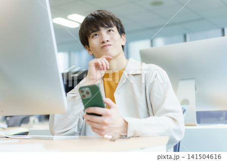 デスクの前に座ってスマホを手に困った顔をしている若い日本人男性 デスクの前に座ってスマホを手に困った顔をしている若い日本人男性 141504616