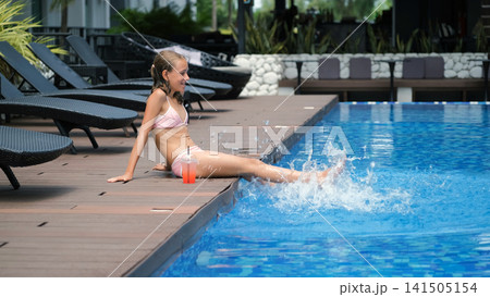 Teenage girl in pink swimsuit sitting on the edge of a blue resort swimming pool swinging her legs in the water with a strawberry cocktail beside her. Leisure summer concept 141505154