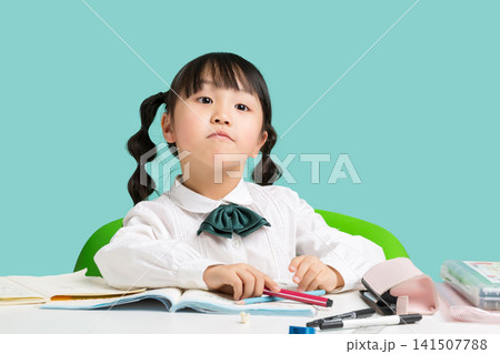 Asian schoolgirl studying with stationery at desk, thinking child looking up on blue background, education concept Asian schoolgirl studying with stationery at desk, thinking child looking up on blue background, education concept 141507788