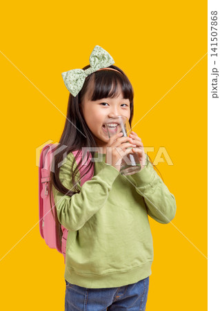 Cute Asian schoolgirl drinking water from glass, healthy hydration concept on yellow background 141507868