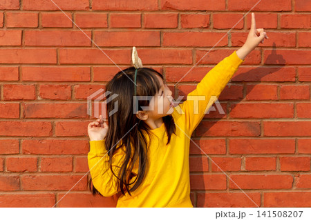 Playful little girl leans against a red brick wall and points her finger high up toward the sky. Playful little girl leans against a red brick wall and points her finger high up toward the sky. 141508207