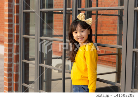 Cute young girl poses next to a large black-framed glass window with a brick wall background. 141508208