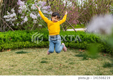 An energetic young girl mid-air jumps with arms raised happily on a park lawn with blooming white blossoms in the background. 141508219
