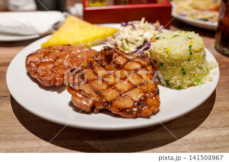 Grilled chicken fillet with grill marks served on a white plate with fried rice, coleslaw, and a side of garlic bread. Delicious restaurant meal served on a wooden table. Healthy protein dish. Grilled chicken fillet with grill marks served on a white plate with fried rice, coleslaw, and a side of garlic bread. Delicious restaurant meal served on a wooden table. Healthy protein dish. 141508967
