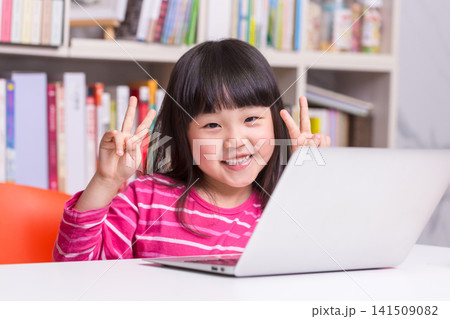 Cheerful Little Asian Girl Making Peace Sign while Studying with Laptop Cheerful Little Asian Girl Making Peace Sign while Studying with Laptop 141509082