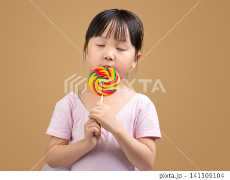 Asian Little Girl Eating Rainbow Lollipop with Eyes Closed on Beige Background Asian Little Girl Eating Rainbow Lollipop with Eyes Closed on Beige Background 141509104
