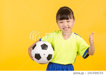 A cheerful East Asian girl soccer player gives a thumbs up while holding a soccer ball against a solid yellow studio wall. A cheerful East Asian girl soccer player gives a thumbs up while holding a soccer ball against a solid yellow studio wall. 141509155