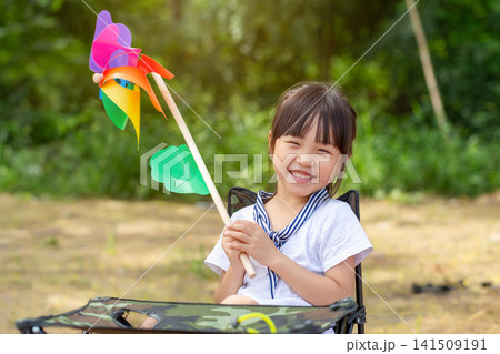 Sweet smiling girl sitting in a camping chair and holding a vibrant rainbow pinwheel outdoors. Sweet smiling girl sitting in a camping chair and holding a vibrant rainbow pinwheel outdoors. 141509191
