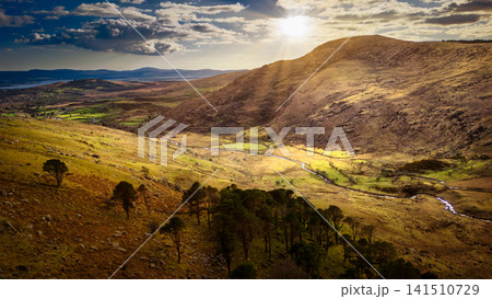 Sunlight streams over a mountain valley with a winding river running through grassy fields and scattered trees under a cloudy sky. Sunlight streams over a mountain valley with a winding river running through grassy fields and scattered trees under a cloudy sky. 141510729
