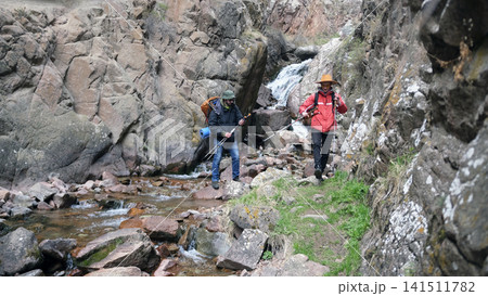Two men are walking along a rocky path next to a stream. A hiking instructor accompanies a tourist on a hike. A trip to the mountains Two men are walking along a rocky path next to a stream. A hiking instructor accompanies a tourist on a hike. A trip to the mountains 141511782