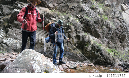 Tourists are walking in nature. Two guys are walking along a rocky path along a stream. A team of hikers crosses a small river Tourists are walking in nature. Two guys are walking along a rocky path along a stream. A team of hikers crosses a small river 141511785
