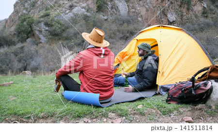 Two men are sitting at a tent in the wilderness. Tourists drink beer and eat sandwiches. They are sitting on a tourist mat Two men are sitting at a tent in the wilderness. Tourists drink beer and eat sandwiches. They are sitting on a tourist mat 141511814