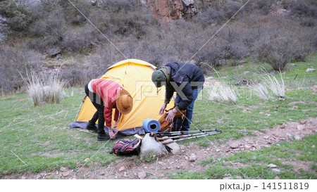 A team of two men sets up a tent on the grass near a mountain gorge. The guys are traveling together 141511819