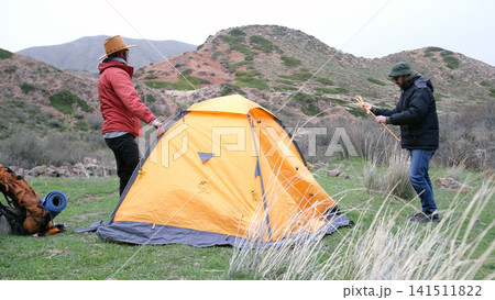 Life in a tent. Hiking in the mountains. Two men are standing next to an orange tent in a grassy field. A trip through the Asian mountains Life in a tent. Hiking in the mountains. Two men are standing next to an orange tent in a grassy field. A trip through the Asian mountains 141511822