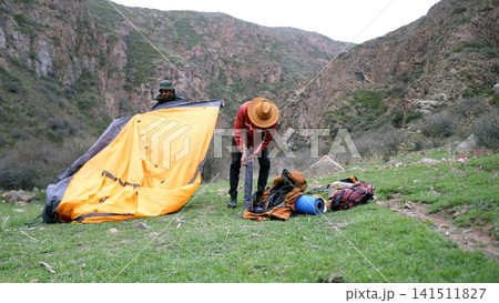 A young tourist puts metal arcs from the tent into a cover. And his partner is folding the canvas of the tent. Two friends travel the world 141511827