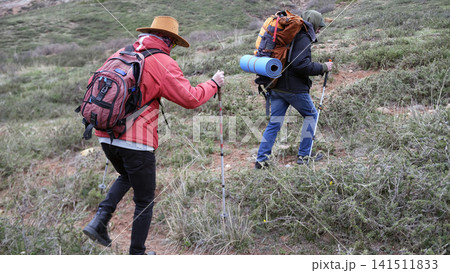 Two tourists with backpacks climb the hill. The climbers are at the beginning of their journey, in a zone of green forests Two tourists with backpacks climb the hill. The climbers are at the beginning of their journey, in a zone of green forests 141511833