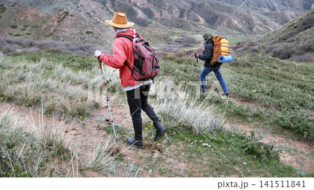 Two students climb the hill with backpacks. Travelers explore new wild areas of the world. Trekking, hiking and tourism. Active pastime Two students climb the hill with backpacks. Travelers explore new wild areas of the world. Trekking, hiking and tourism. Active pastime 141511841