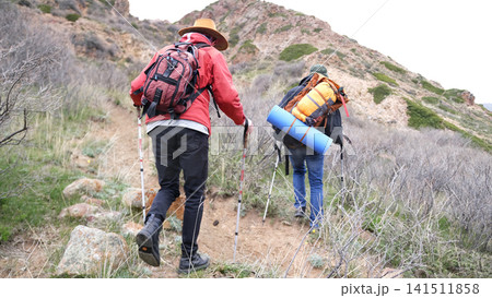 Two travelers climb the mountain with backpacks and trekking poles. The guide is taking his friend camping all weekend Two travelers climb the mountain with backpacks and trekking poles. The guide is taking his friend camping all weekend 141511858