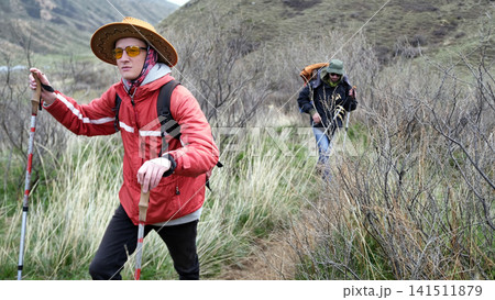 A man in a red jacket, yellow glasses and a cowboy hat walks along the trail with trekking poles. He is followed by his friend A man in a red jacket, yellow glasses and a cowboy hat walks along the trail with trekking poles. He is followed by his friend 141511879