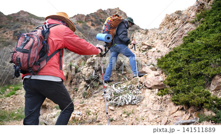 Tourists - climbers climb the mountain. Extreme and freedom. Two friends are walking up a mountain path past rocks and juniper bushes Tourists - climbers climb the mountain. Extreme and freedom. Two friends are walking up a mountain path past rocks and juniper bushes 141511889