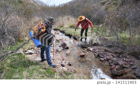 A professional guide watches as his ward tourist crosses the river. A couple of friends are trekking in the mountains. Unity with nature A professional guide watches as his ward tourist crosses the river. A couple of friends are trekking in the mountains. Unity with nature 141511903