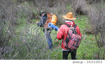 A guy in a cowboy hat and a red jacket is following his friend with a hiking backpack. Weekend camping trip. Active pastime A guy in a cowboy hat and a red jacket is following his friend with a hiking backpack. Weekend camping trip. Active pastime 141511915