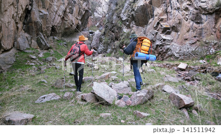 Two wanderers walk to the waterfall along a rocky gorge. Weekend camping trip. Tourists are hiking in the highlands, they have backpacks Two wanderers walk to the waterfall along a rocky gorge. Weekend camping trip. Tourists are hiking in the highlands, they have backpacks 141511942