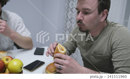 Close-up of a man eating a sandwich with cheese and sausage, and thinking about something. A simple breakfast for two cute gay men Close-up of a man eating a sandwich with cheese and sausage, and thinking about something. A simple breakfast for two cute gay men 141511960