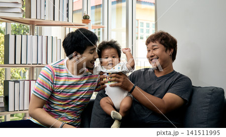 Two smiling men holding a baby on a sofa at home, sharing happiness, care, affection, bonding, support, comfort, love, and joyful togetherness in a bright living room Two smiling men holding a baby on a sofa at home, sharing happiness, care, affection, bonding, support, comfort, love, and joyful togetherness in a bright living room 141511995