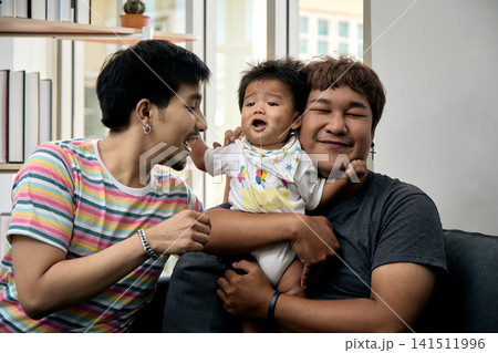 Two men comforting a crying baby on a sofa at home, showing care, affection, support, tenderness, bonding, comfort, concern, caregiving, and emotional family connection 141511996