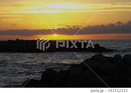 Sunset over calm ocean waves, with rocky shoreline silhouetted against vibrant orange and yellow sky, creating a serene and tranquil evening atmosphere by the water Sunset over calm ocean waves, with rocky shoreline silhouetted against vibrant orange and yellow sky, creating a serene and tranquil evening atmosphere by the water 141512315
